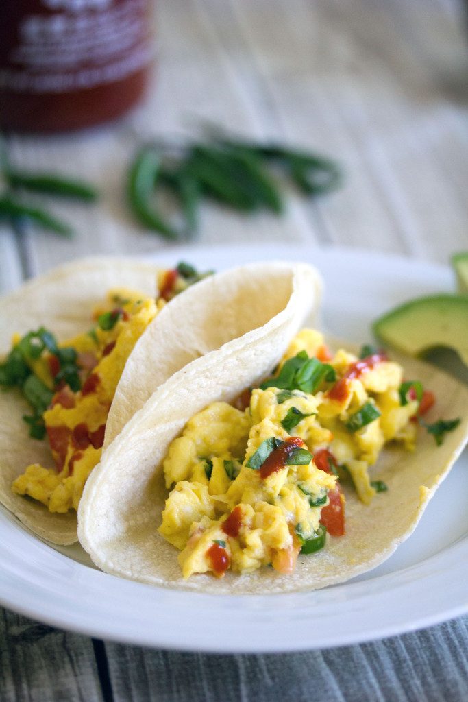 Head-on closeup view of two Thai scrambled egg tacos with basil and Sriracha and sliced avocado on a white plate with Thai peppers and Sriracha bottle in background