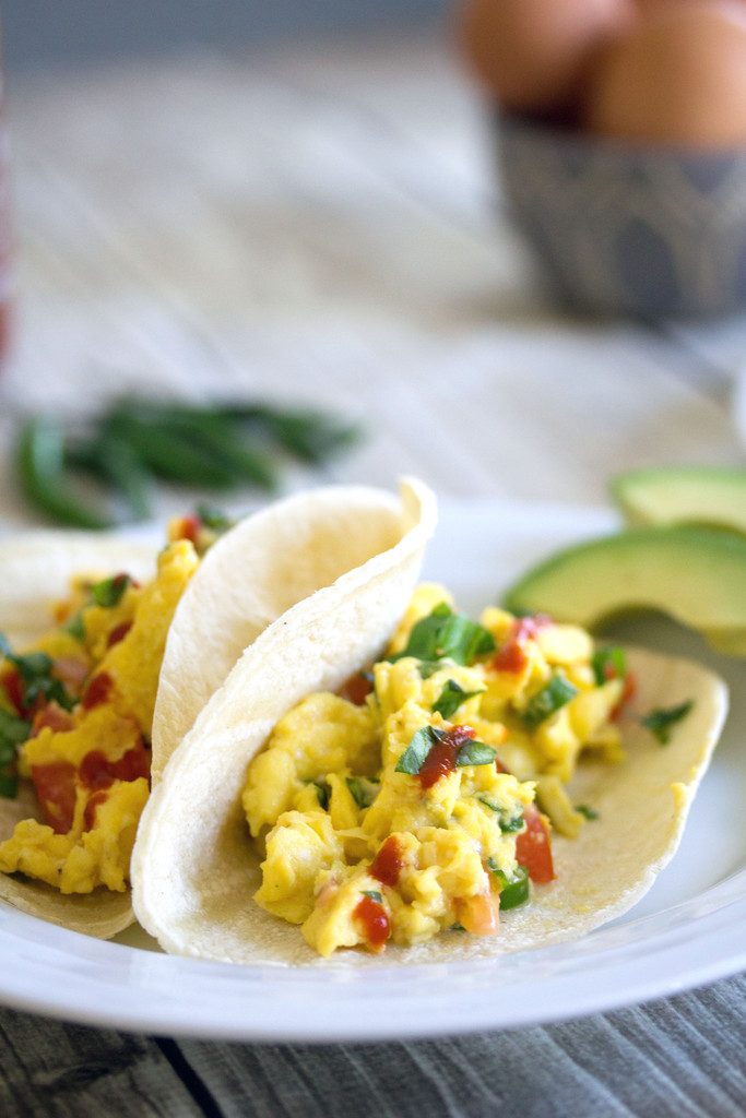 Head on view of two Thai scrambled egg tacos drizzled with Sriracha in flour tortillas on white plate with avocado, Thai peppers, and eggs in the background