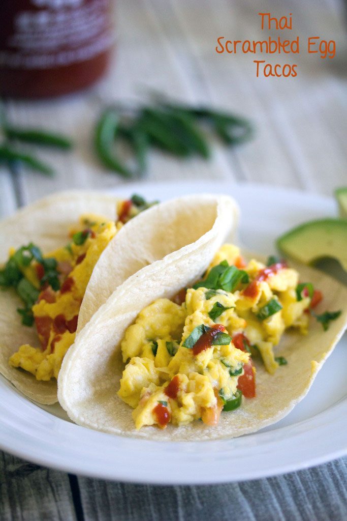 Head-on view of Thai Scrambled Egg Tacos with Thai peppers and Sriracha on a white plate with sliced avocado and more peppers in the background with recipe title at top