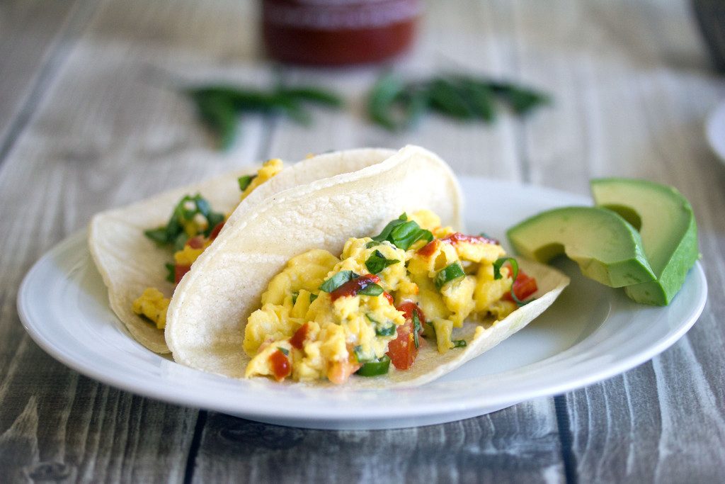 Landscape view of two Thai scrambled egg tacos topped with basil and Sriracha on a white plate with avocado, Thai peppers, and Sriracha bottle in the background