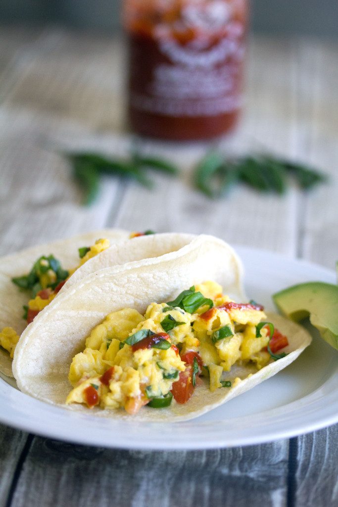 Head-on view of Thai Scrambled Egg Tacos with Thai peppers and Sriracha on a white plate with sliced avocado and more peppers in the background