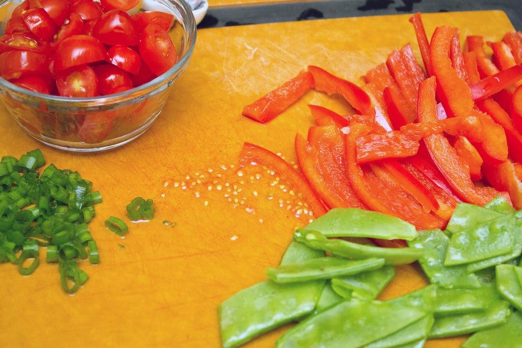 Overhead view of cutting board with chopped peppers, pea pods, scallions, and tomatoes