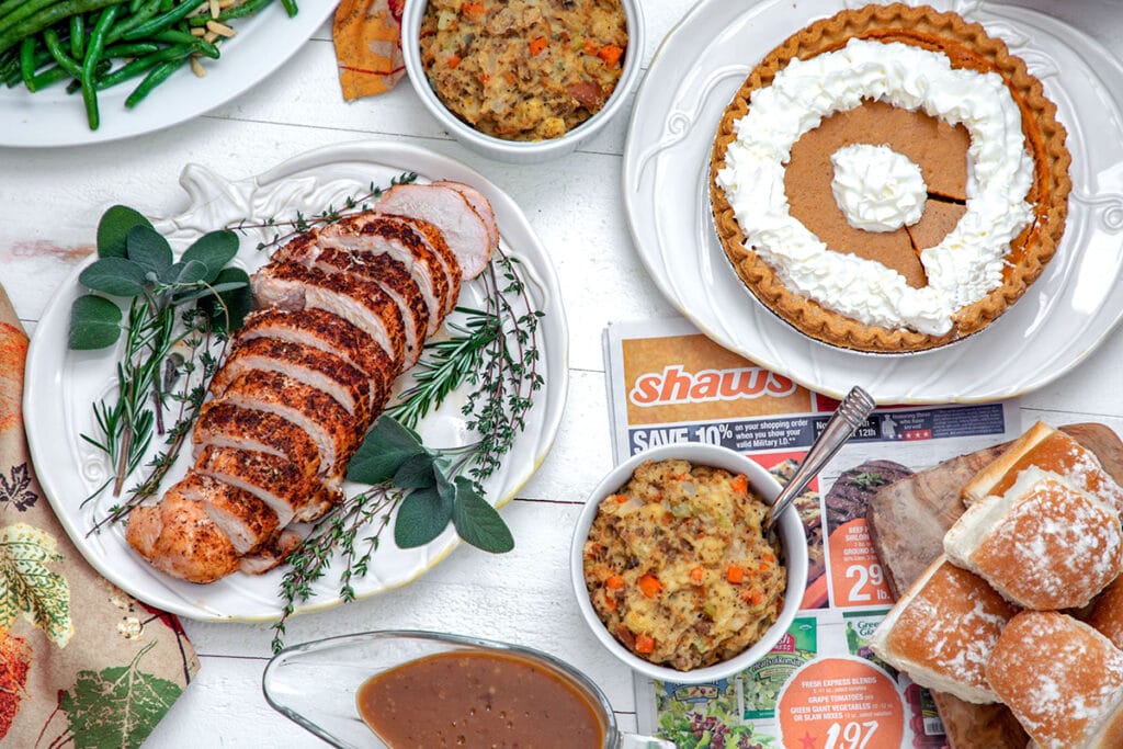 Landscape view of full Thanksgiving dinner, including turkey breast on platter, stuffing, pumpkin pue with whipped cream, dinner rolls, green beans, and gravy.