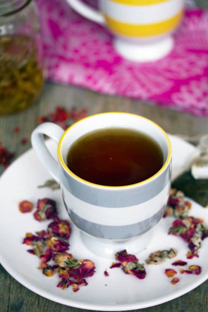 Close-up view of English rose cocktail in a striped teacup on a white plate surrounded by dried rosebuds and teabag and jar of simple syrup in the background