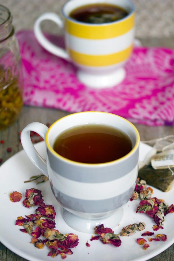 Head-on photo of two striped teacups filled with English Rose cocktail with one on a white plate, surrounded by dried roses and a tea bag