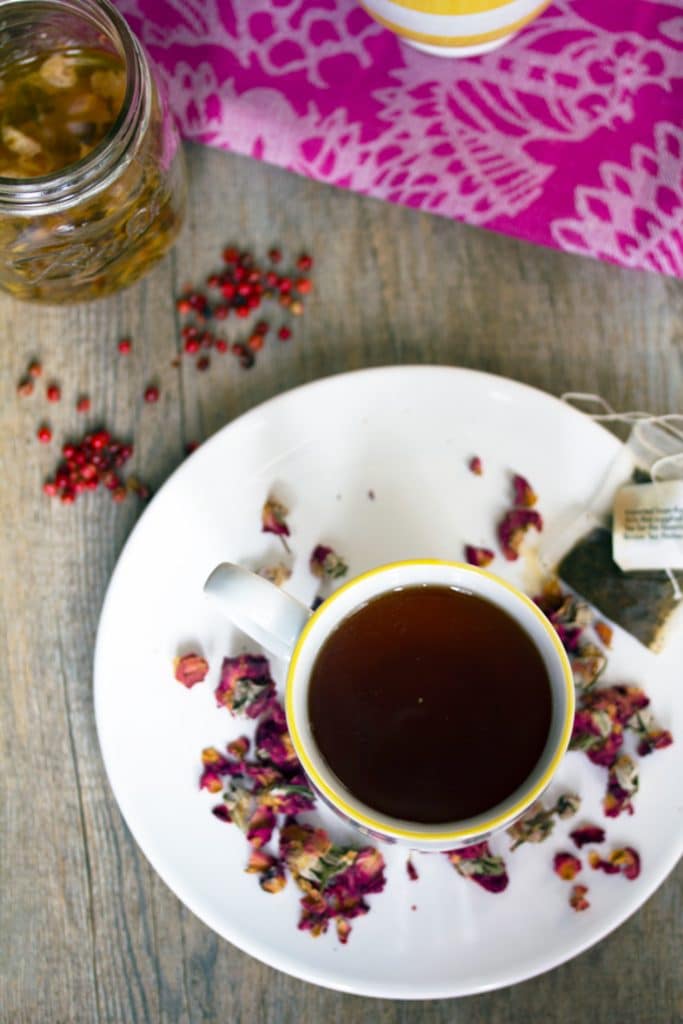 Overhead view of English rose cocktail on a white plate with dried rosebuds and a tea bag with pink peppercorns around plate and a jar of simple syrup in the background