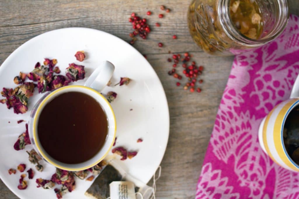 Overhead view of English rose cocktail in a teacup on a white plate surrounded by dried rosebuds with pink peppercorns, a teabag, and jar of simple syrup