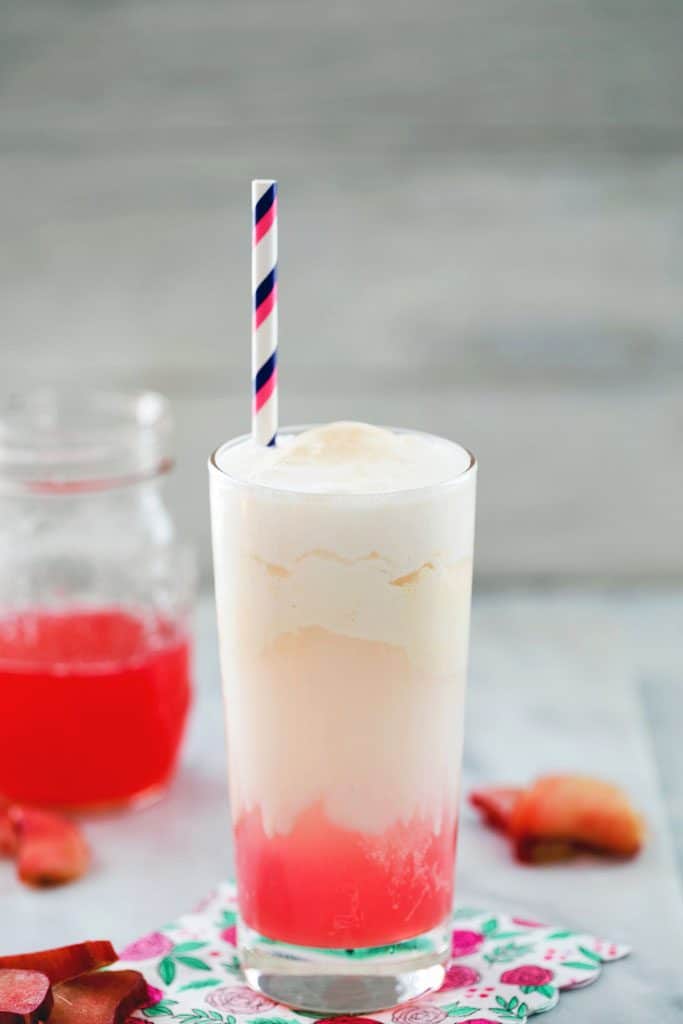 View of rhubarb ice cream soda with hot pink syrup at the bottom of the glass and a striped straw, with jar of pink syrup and chopped rhubarb in the background