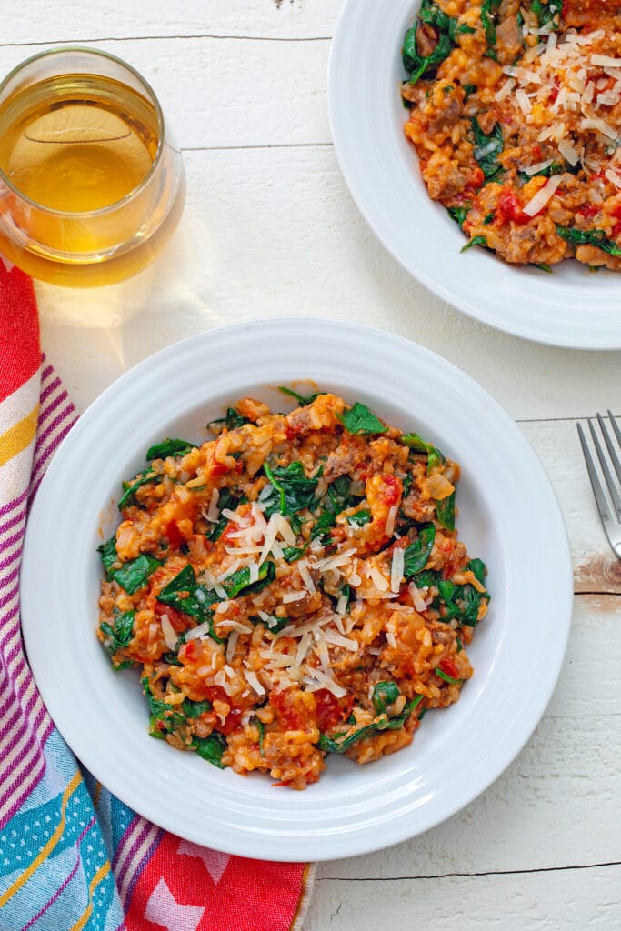 Tomato and sausage risotto with spinach in a bowl with a second bowl and glass of white wine in background and fork on the side