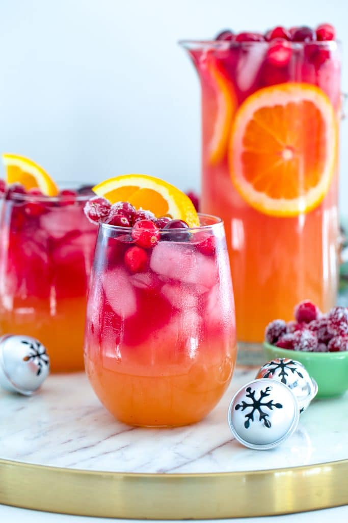 Head-on view of glass of tropical cranberry sangria with second glass, pitcher, mini holiday bells, and bowl of sugared cranberries in the background