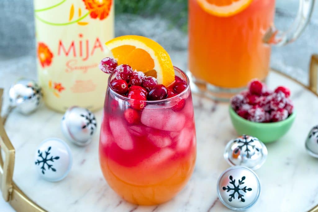 Landscape overhead view of glass of tropical cranberry sangria on a gold and marble surface surrounded by bottle of white Mija Sangria, mini holiday bells, and sugared cranberries