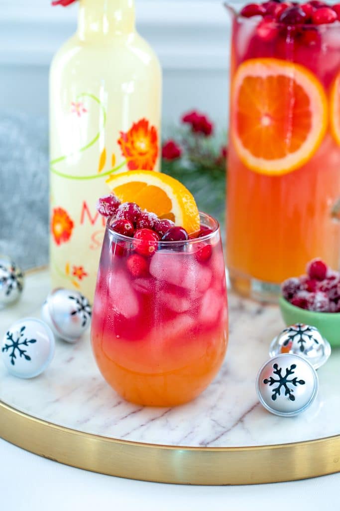 Head-on view of glass of tropical cranberry sangria, with bottle of white Mija sangria, pitcher of sangria, mini holiday bells, and bowl of sugared cranberries in the background