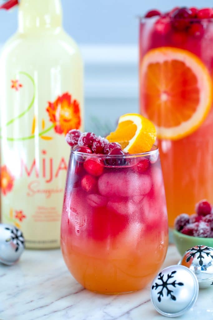 Head-on closeup view of glass of tropical cranberry sangria with bottle of white Mija sangria, pitcher of sangria and mini holiday bells in the background