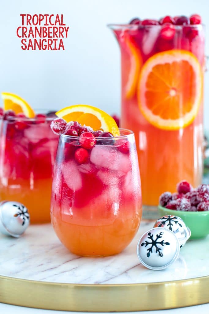 Head-on view of glass of tropical cranberry sangria on a marble and gold surface with a second glass, a pitcher of cocktail, sugared cranberries, and mini holiday bells in the background with recipe title at top of image
