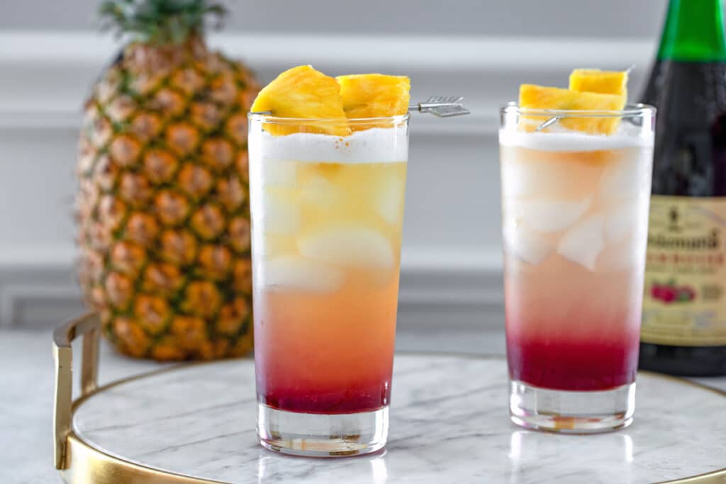 Landscape head-on view of two tropical gin sunrise cocktails on a marble tray with whole pineapple and bottle of raspberry lambic in background