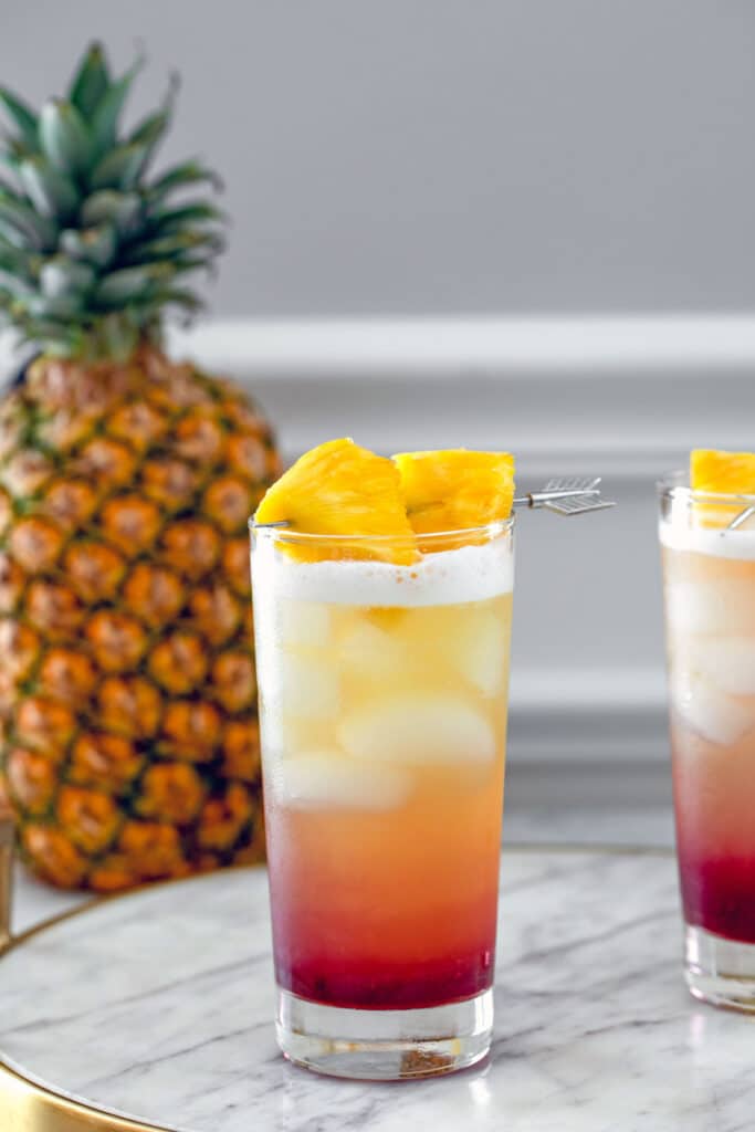 Head-on view of tropical gin sunrise cocktail on a marble tray with pineapple garnish and whole pineapple in the background