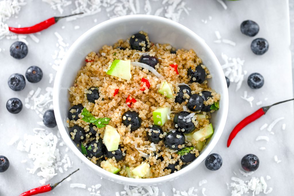 Landscape overhead view of a white bowl of tropical quinoa salad on a marble surface with shredded coconut, blueberries, and red chili peppers all around