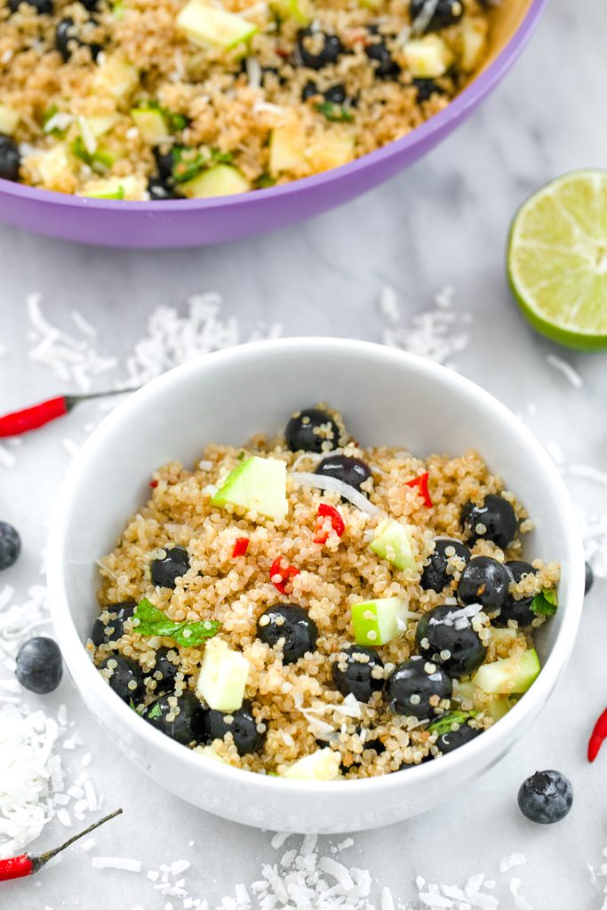 Overhead view of a white bowl of a tropical quinoa salad with blueberries, green apple, coconut, and red chili peppers on a marble surface with larger bowl in the background and coconut, chili peppers, and blueberries scattered around