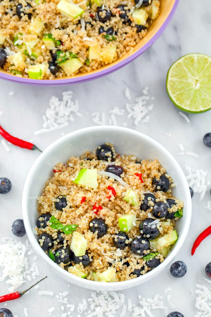 Bird's eye view of a white bowl of tropical quinoa salad on a marble surface with shredded coconut, blueberries, red chili peppers, a lime half, and large serving bowl of salad in background