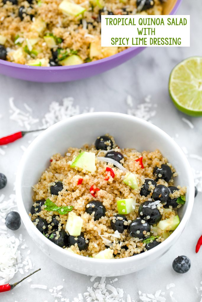 Overhead view of a white bowl of a tropical quinoa salad with blueberries, green apple, coconut, and red chili peppers on a marble surface with larger bowl in the background, coconut, chili peppers, and blueberries scattered around and recipe title at top