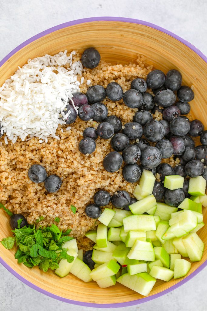 Overhead view of a serving bowl filled with quinoa, blueberries, shredded coconut, chopped green apples, and chopped mint before being tossed