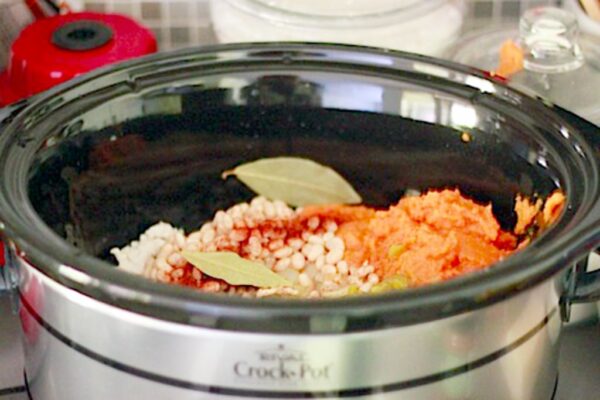 Landscape photo of slow cooker filed with chili ingredients, including white beans, pumpkin, chili powder, and bay leaves