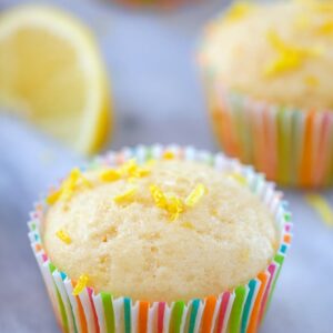 Head-on view of lemon muffin topped with lemon zest in a colorful muffin liner with a second muffin and lemon wedge in the background and "Tuscan Lemon Muffins" text at top