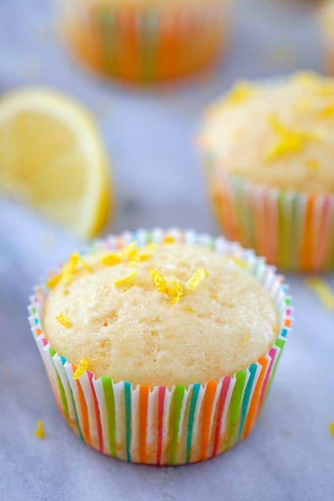 Head-on view of lemon muffin topped with lemon zest in a colorful muffin liner with a second muffin and lemon wedge in the background