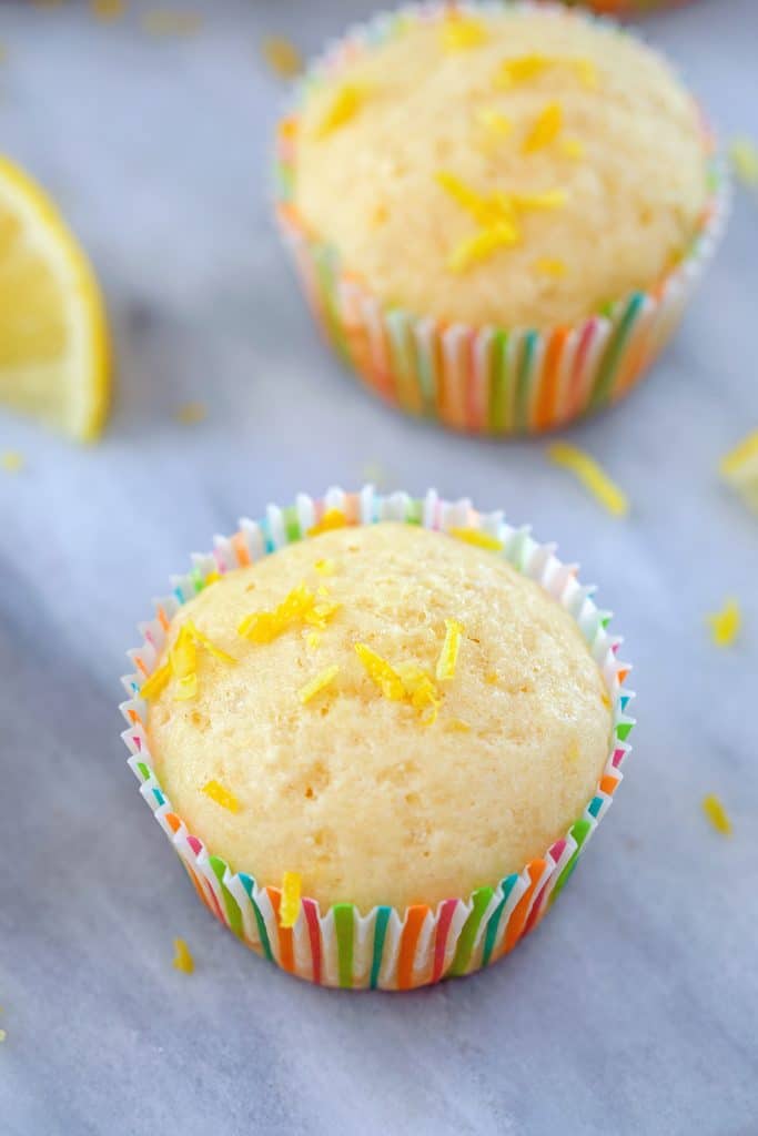 Two Tuscan lemon muffins on a marble surface with lemon zest scattered around and a lemon wedge in the background