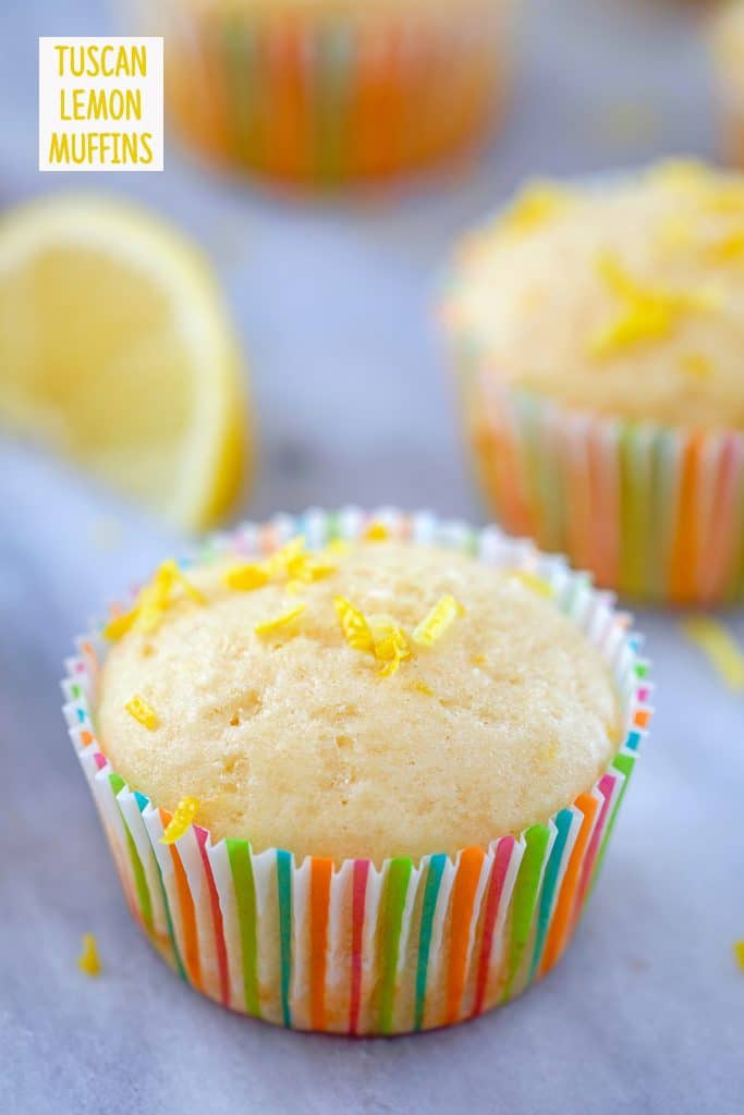 Head-on view of lemon muffin topped with lemon zest in a colorful muffin liner with a second muffin and lemon wedge in the background and "Tuscan Lemon Muffins" text at top