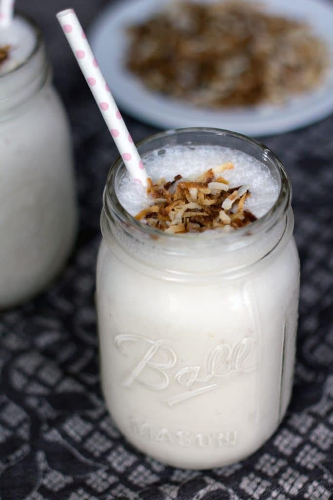 Close up of vanilla coconut milkshake in a mason jar topped with toasted coconut and a pink polka dot straw with a plate of toasted coconut in the background