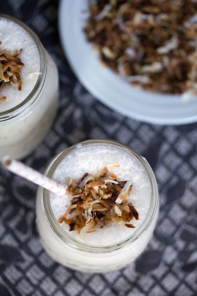 Overhead view of two vanilla coconut milkshakes topped with toasted coconut and straws with a plate of toasted coconut in the background