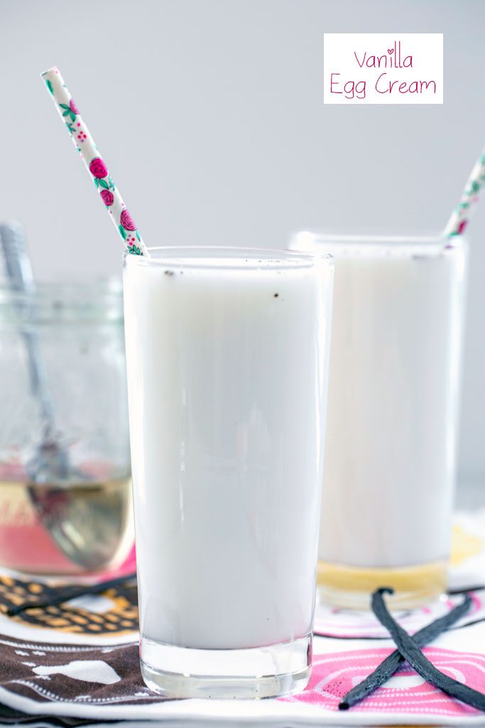 Head-on view of a tall glass of vanilla egg cream with a second vanilla egg cream, jar of simple syrup, and vanilla beans in the background with recipe title at top