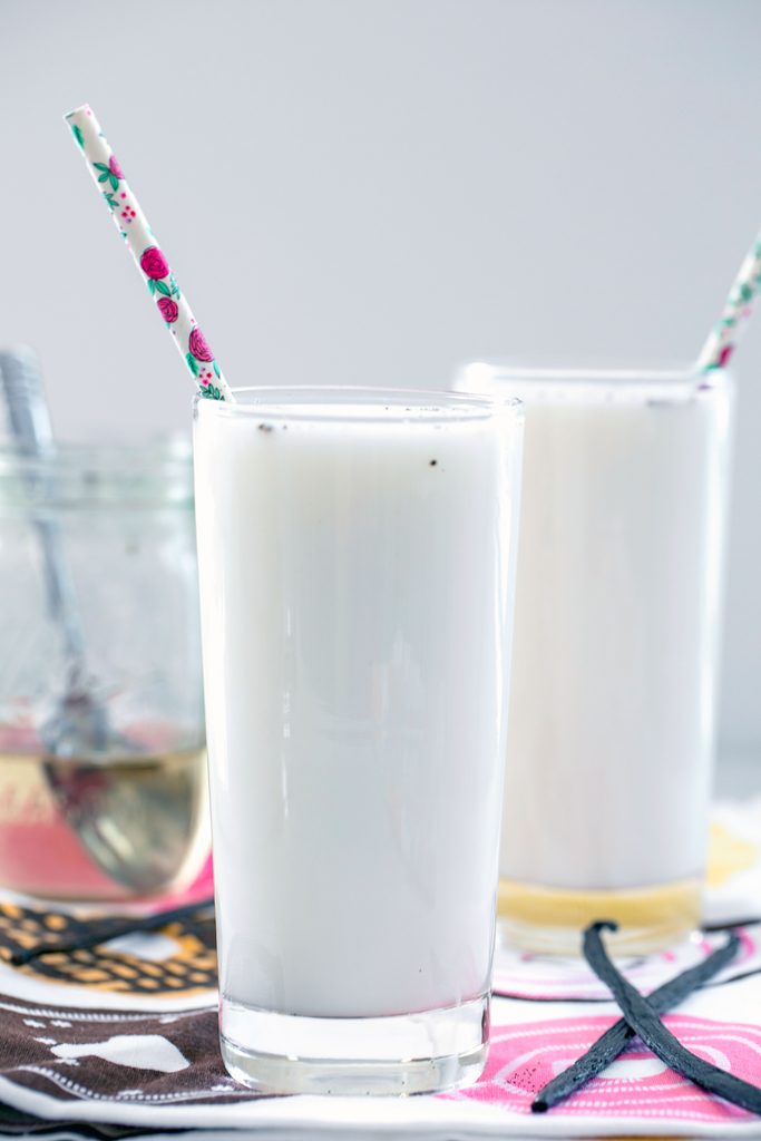 Head-on view of a tall glass of vanilla egg cream with a second vanilla egg cream, jar of simple syrup, and vanilla beans in the background