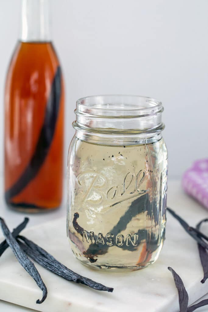 Head-on view of a mason jar with vanilla syrup with vanilla beans all around and bottle of vanilla extract in background.