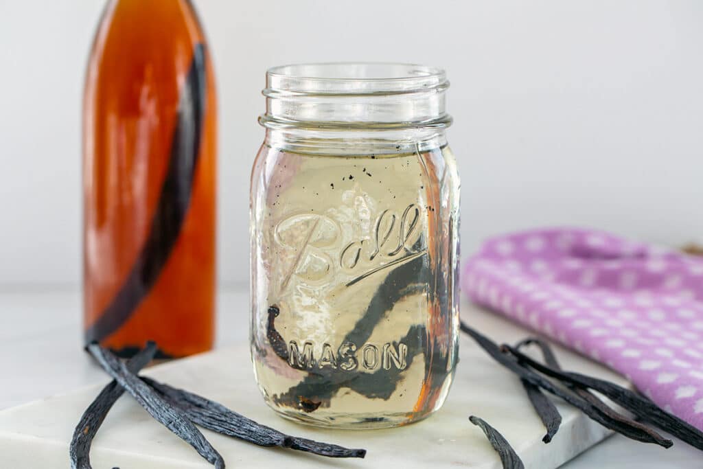 Landscape head-on view of a mason jar of vanilla syrup with bottle of vanilla extract in background and vanilla beans all around.