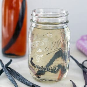 Head-on view of a mason jar with vanilla syrup with vanilla beans all around and bottle of vanilla extract in background.