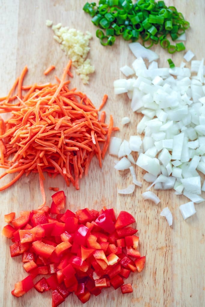 Overhead view of a wooden cutting board with chopped red peppers, shredded carrots, chopped onion, minced garlic, and chopped scallions