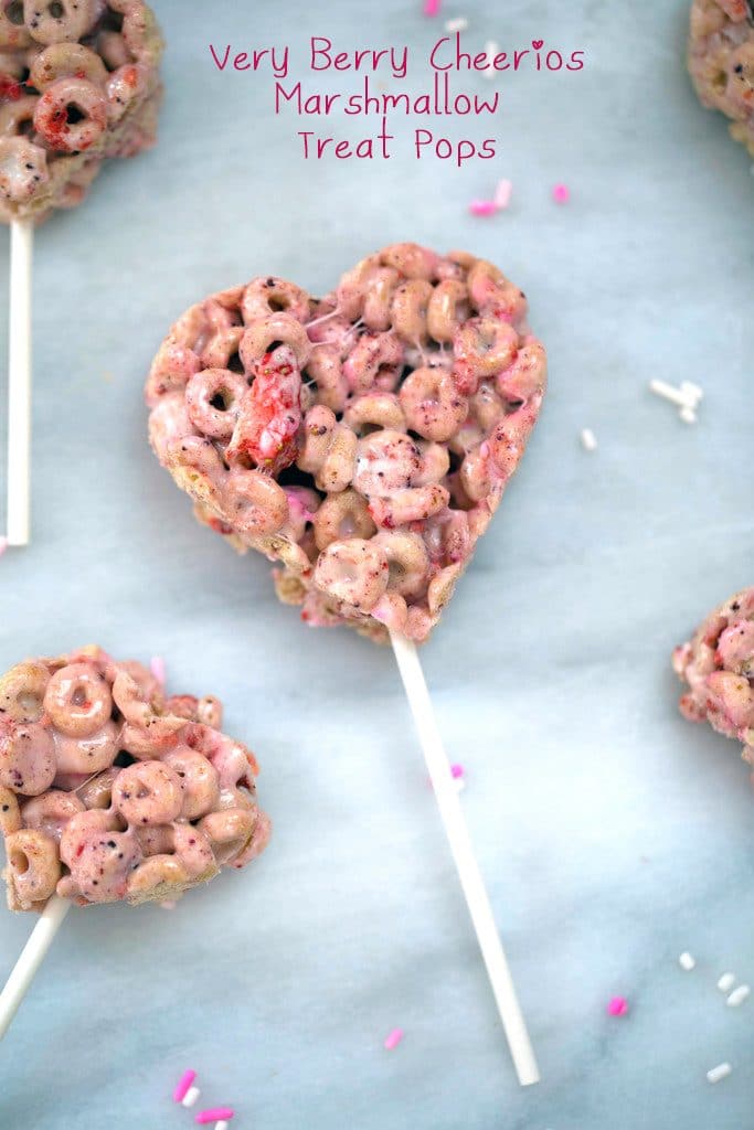 Overhead view of pink heart-shaped Very Berry Cheerios Treats on a popsicle stick on a marble surface with recipe title at top