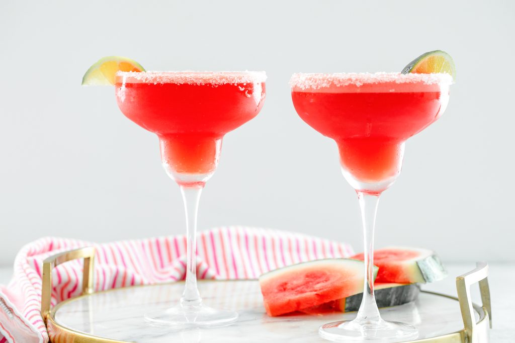 Landscape head-on view of two watermelon margaritas with sea salt rims and lime wedge garnishes on a marble tray with watermelon slices also on tray