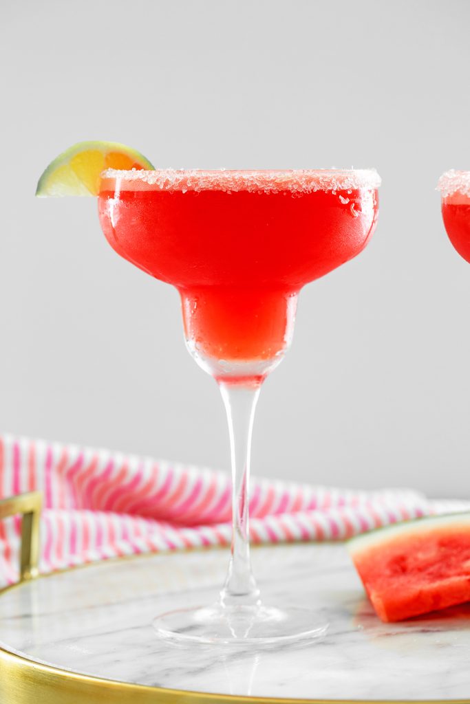 Head-on view of watermelon margarita with sea salt rim and lime wedge with watermelon slice and second margarita in background