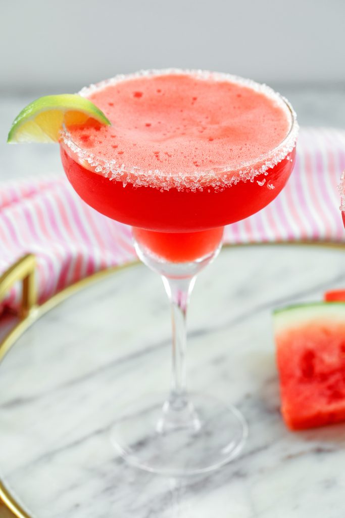 Angled view of watermelon margarita with sea salt rim and lime wedge garnish on a marble tray with watermelon slices in background