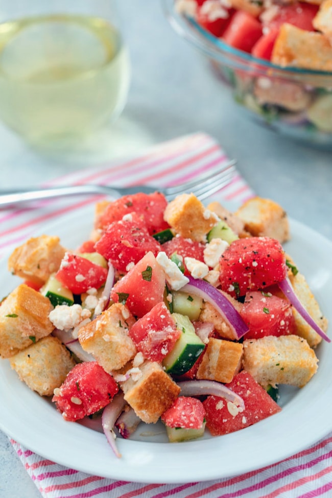 A head-on view of watermelon panzanella salad on a white plate and pink striped towel with big bowl of salad, fork, and glass of white wine in the background