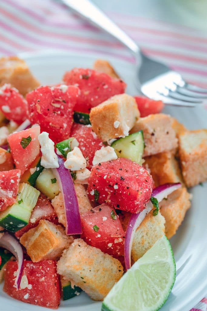 Close-up view of watermelon panzanella salad, including watermelon, red onion, cubed bread, cucumber, feta cheese, and a lime wedge