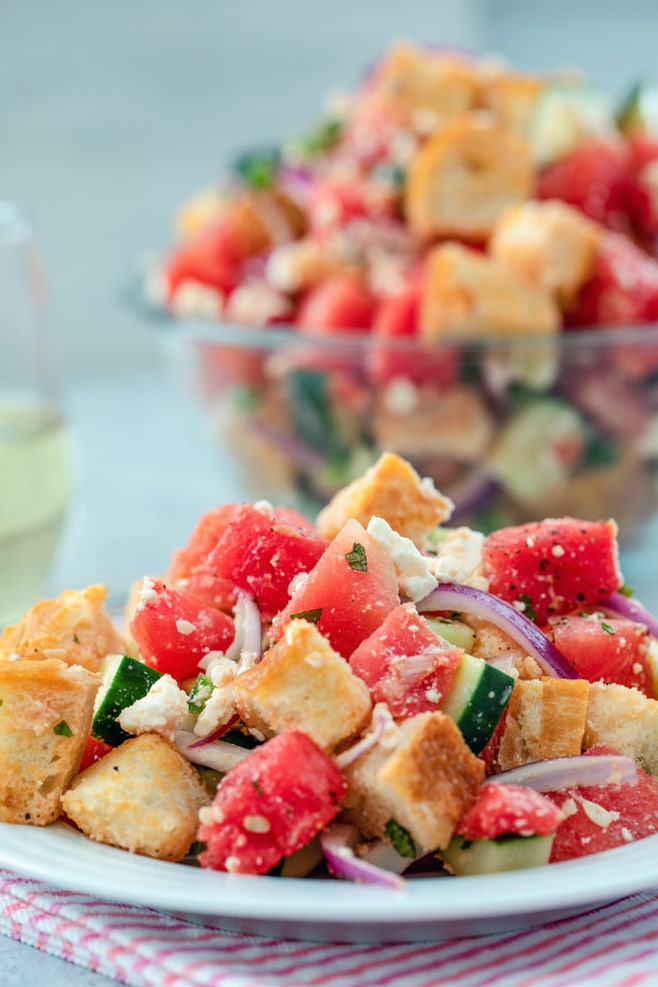 Head on view of watermelon panzanella salad piled on a white plate with a heaping bowl of it in the background, along with a glass of white wine