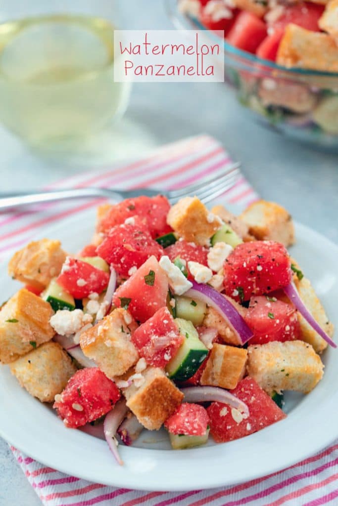 A head-on view of watermelon panzanella salad on a white plate and pink striped towel with big bowl of salad, fork, and glass of white wine in the background and "Watermelon Panzanella" text at top