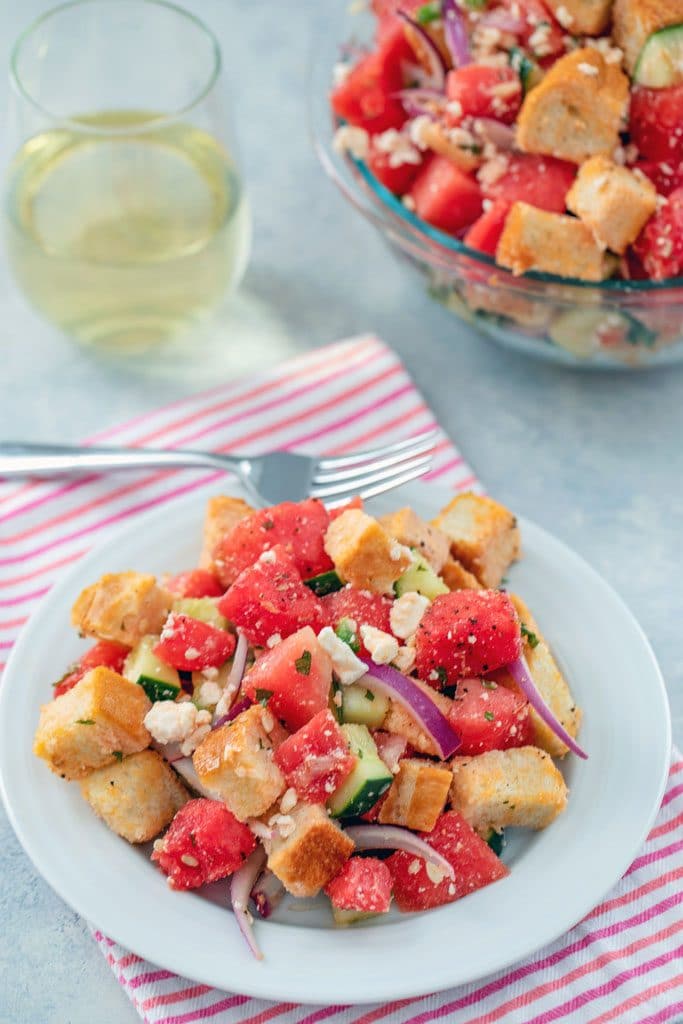 Bird's eye view of watermelon panzanella on a white plate and pink striped towel with a fork, glass of white wine, and large bowl of salad in the background