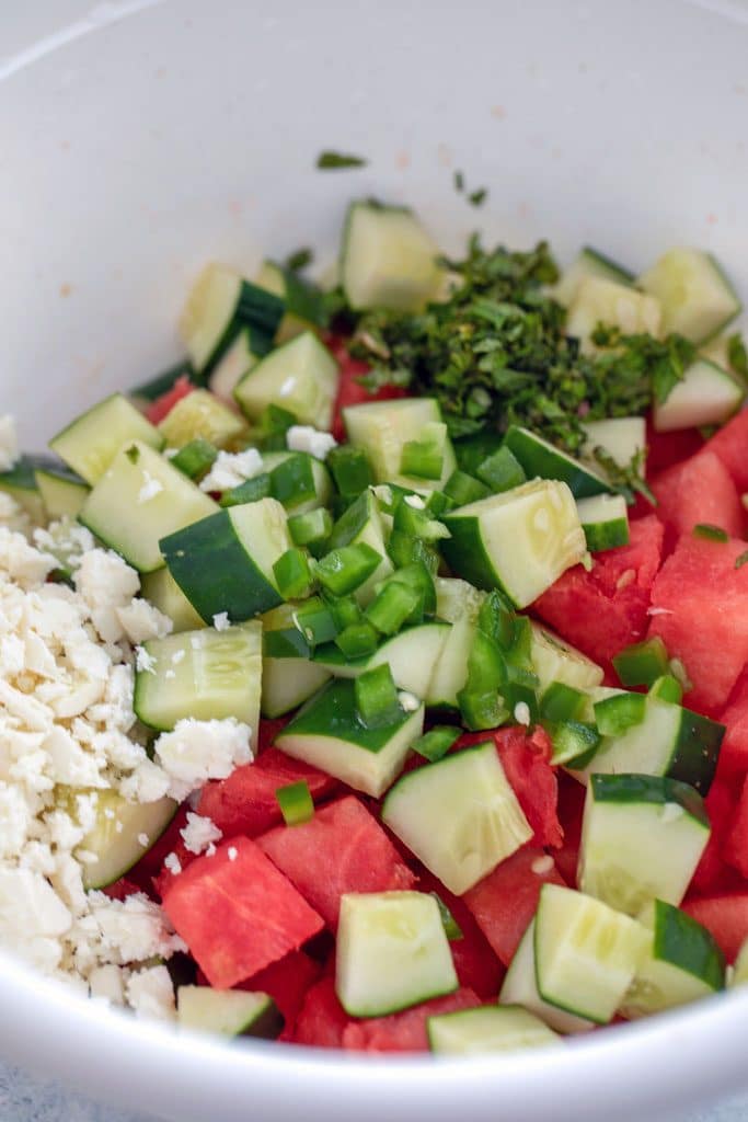 A big bowl filled with cubed watermelon, cubed cucumbers, feta cheese, diced jalapeño pepper, and chopped mint.