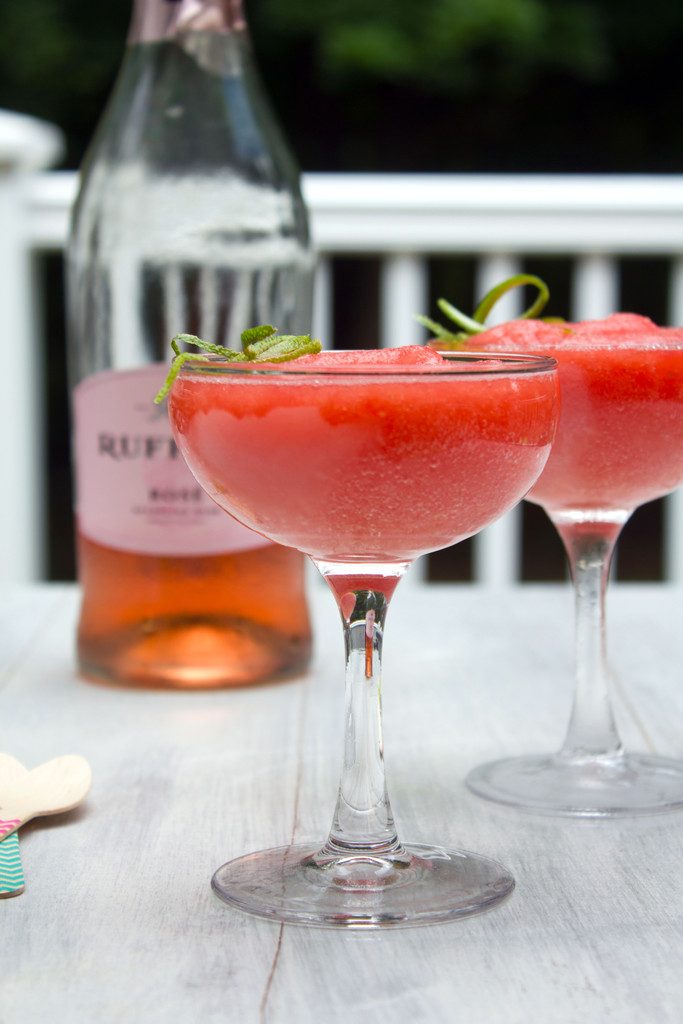 Head-on view of two coup glasses of watermelon rosé slushies on a table outside with bottle of rosé in the background