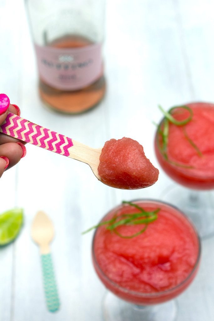 Overhead view of coup glasses with watermelon rosé sangria with a hand holding a wooden spoon with a scoop of slushy on it and a bottle of rosé in the backgroudnd